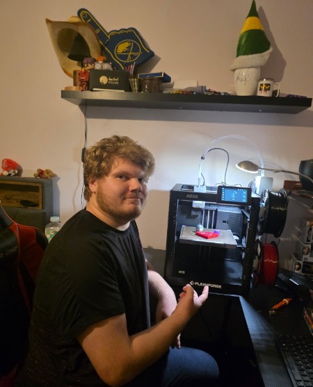 Owner of Plouffe 3D Prints seated at the workbench beside a Flashforge 3D printer running a pink print