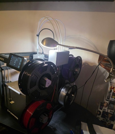 Filament spool rack loaded with black, grey and red filament beside a 3D printer and task lamp